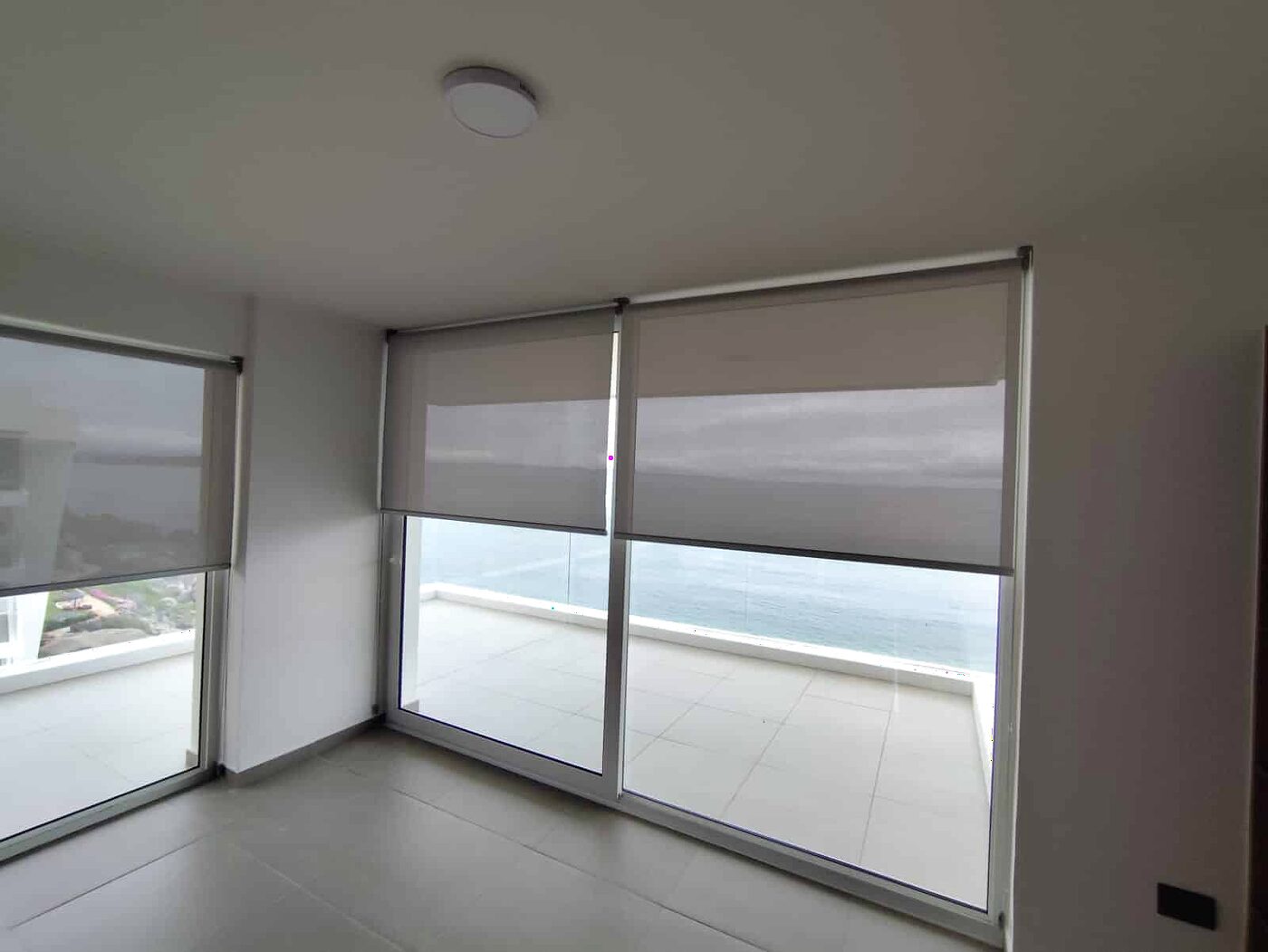 Cortinas roller sunscreen sobre el ventanal de un living con salida a terraza y vista al mar