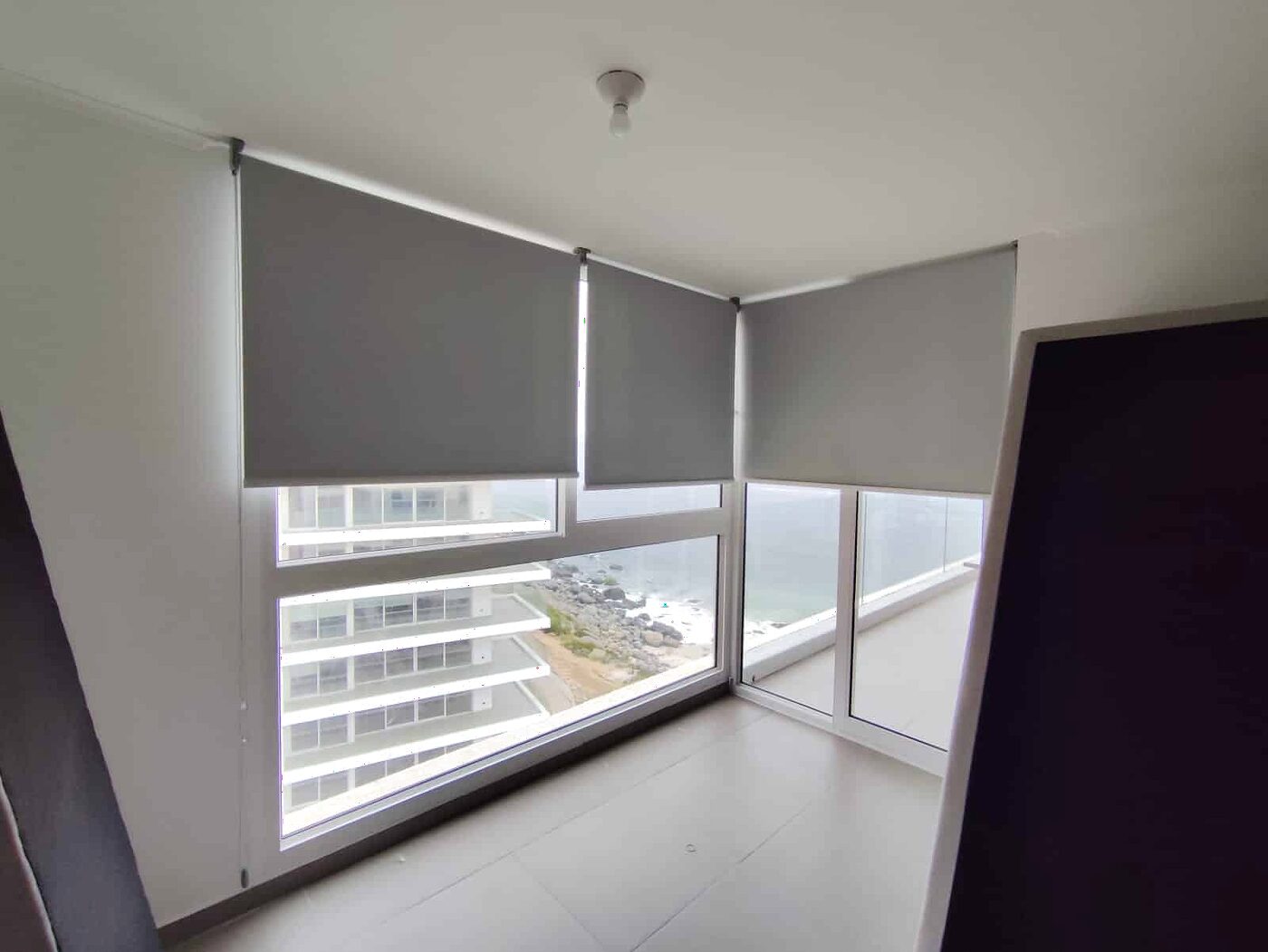 Cortina roller gris en la ventana esquina de un dormitorio, con vista a los edificios