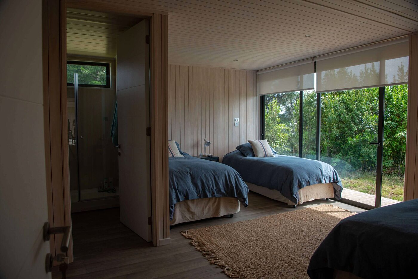 Dormitorio de dos camas con revestimiento de madera clara y ventanal al bosque